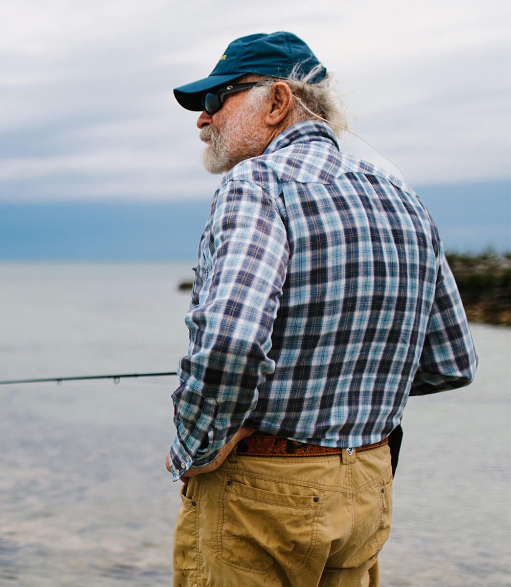 Flip Pallott's back is towards the camera and he is holding onto a fly fishing rod. There is water in the background. 